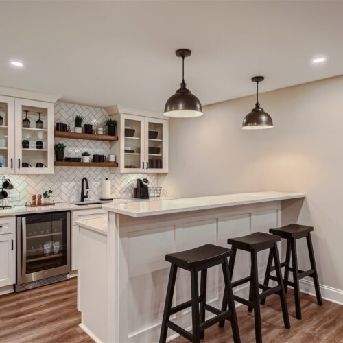 A modern kitchen with a white color scheme. It features a bar area with three black wooden bar stools, white countertops, a sink, glass-front cabinets, a built-in wine fridge, and pendant lights. The backsplash is tiled in a herringbone pattern. General Contractor and Home Remodeler serving Atlanta, Alpharetta, Marietta, Decatur, Cumming, Johns Creek
