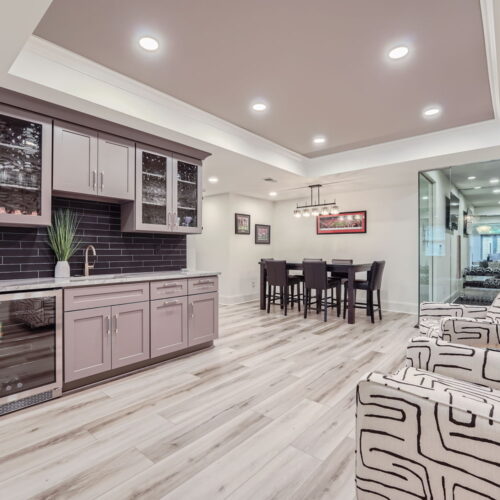Modern Lange Basement with open layout featuring a wet bar with gray cabinets, glass doors, and a wine cooler. A tall dining table with chairs is in the background. Two patterned armchairs are positioned in the foreground, and recessed lighting brightens the space.
